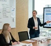 Das Bild zeigt einen Englischkurs für ein Vertriebsteam in einem modernen Frankfurter Büro. Das Meeting findet in einem zeitgemäßen Konferenzraum mit hellem Holzboden, holzvertäfelten Akustikwänden und einem großen Fenster mit freiem Blick auf die markante Frankfurter Skyline statt. Die Kursleiterin steht vorne im Raum und lenkt die Aufmerksamkeit des Teams auf einen großen Bildschirm,  auf dem eine Präsentation mit dem Titel „FRANKFURT VERTRIEBSSTRATEGIE & MARKTWACHSTUM“ gezeigt wird.
