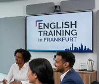 Das Bild zeigt eine Schulung für Wirtschaftsenglisch, die in Frankfurt am Main stattfindet. Große, bodentiefe Fenster hinter der Gruppe bieten einen Panoramablick auf die Frankfurter Skyline. Ein mobiles Whiteboard zeigt handgezeichnete Diagramme und Texte, die sich thematisch um „BUSINESS ENGLISH COMMUNICATION“ drehen. Pfeile in Form einer Mindmap verweisen auf Themen wie „ENGLISH TRAINING FRANKFURT“, „EFFECTIVE PRESENTATIONS“, „MEETINGS“, „SKILLS“ und „VOCABULARY“.