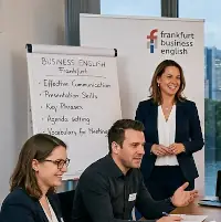 Das Bild zeigt einen professionellen Kurs für Wirtschaftsenglisch – Grammatik und schriftlicher Ausdruck – in Frankfurt am Main. Schauplatz ist ein moderner Konferenz- oder Schulungsraum eines Unternehmens am Abend. Ein riesiges, vom Boden bis zur Decke reichendes Fenster eröffnet einen weiten Blick auf die Frankfurter Skyline, die sich vor dem Dämmerungshimmel hell erleuchtet abzeichnet, während der Main an den Wolkenkratzern vorbeifließt.