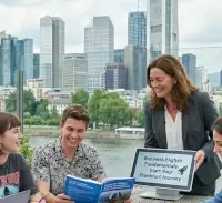 Die Szene spielt auf einer modernen Außenterrasse mit direktem Blick auf den Main und die Frankfurter Skyline – einschließlich markanter Wolkenkratzer wie dem Commerzbank Tower. Ein Glasschild auf einer steinernen Säule zur Linken weist den Ort als „YOUNG ADULT ENGLISH PROGRAM: FRANKFURT“ aus, versehen mit dem Slogan „Global Communications for the Next Generation“. Das Wetter wirkt hell und klar und verleiht der Unterrichtsstunde eine lebendige, urbane Atmosphäre.